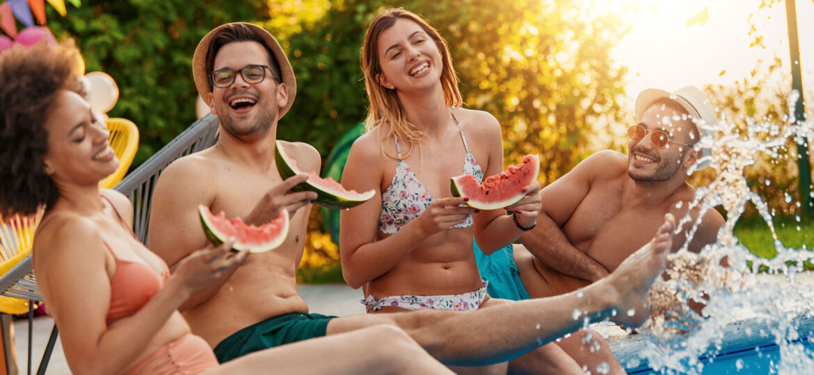 Group of 4 friends sitting on the edge of the pool splashing water with their feed. They are wearing bathing suits, laughing and smiling, eating watermelon and having a good time. There is a colorful banner in the background indicating this is a party on a warm summer day. Heliocol makes swimming enjoyable for more of the year!
