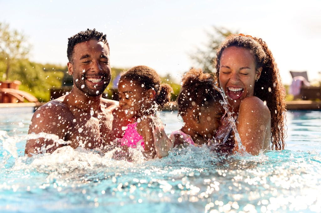 A family of 4 plays in the pool. The husband and wife each hold one of their little girls in their arms. There eyes are closed and they smile wide as the girls splash there arms with joy. Behind them you can see some poolside furniture and a clear blue sky.