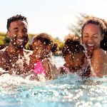 A family of 4 plays in the pool. The husband and wife each hold one of their little girls in their arms. There eyes are closed and they smile wide as the girls splash there arms with joy. Behind them you can see some poolside furniture and a clear blue sky.