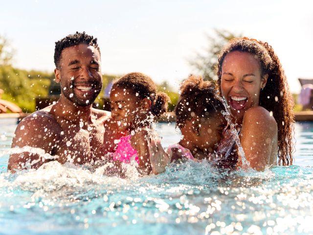 A family of 4 plays in the pool. The husband and wife each hold one of their little girls in their arms. There eyes are closed and they smile wide as the girls splash there arms with joy. Behind them you can see some poolside furniture and a clear blue sky.