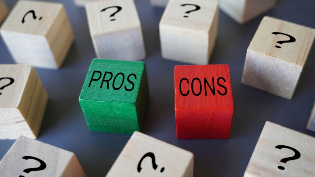 Wooden blocks with "PROS" and "CONS" written on them in black letters, the "PROS" block is green and the "CONS" block is red. Surrounding these blocks are other wooden blocks with black question marks on them. The image conveys a concept of weighing pros and cons amidst uncertainty such as when comparing different types of pool heating systems.