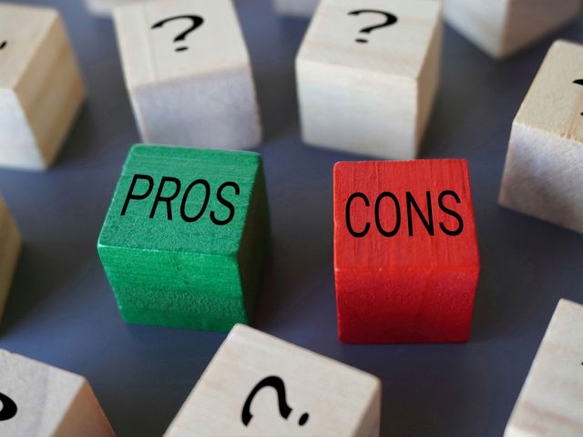 Wooden blocks with "PROS" and "CONS" written on them in black letters, the "PROS" block is green and the "CONS" block is red. Surrounding these blocks are other wooden blocks with black question marks on them. The image conveys a concept of weighing pros and cons amidst uncertainty such as when comparing different types of pool heating systems.