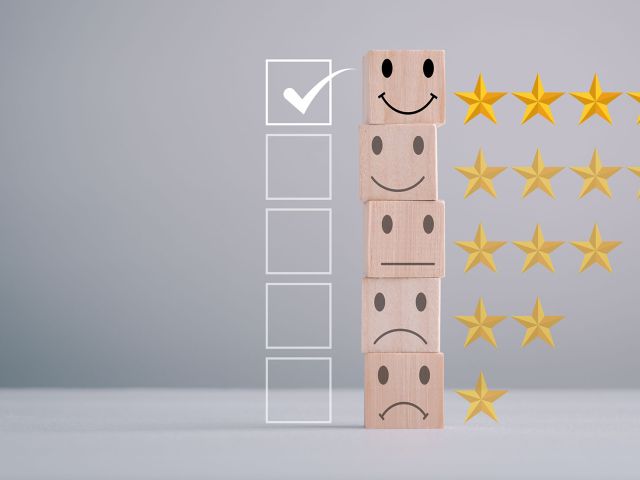 A stack of wooden blocks displaying different facial expressions, from top to bottom: a smiling face with five gold stars, a neutral face with four gold stars, a slightly sad face with three gold stars, and a very sad face with one gold star. To the left of the stack is a vertical row of empty checkboxes, with the topmost box checked. The image represents a rating or feedback system. This is symbolic of the Heliocol Solar Pool Heating System being the top choice for our customers.