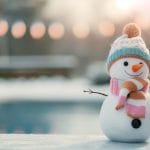 A cheerful snowman with a carrot nose and a smile stands outdoors on a snowy surface. It is adorned with a pastel-colored striped scarf and a knitted beanie featuring a pom-pom. The background is softly blurred, showing a wintery scene with a pool and warm lights, creating a cozy and festive atmosphere.