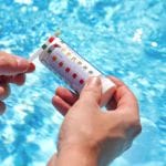 A person holds a pool test strip and a container with a color-coded chart in front of a sparkling blue swimming pool. The test strip is being compared to the chart, likely to assess water quality parameters such as pH and chlorine levels.