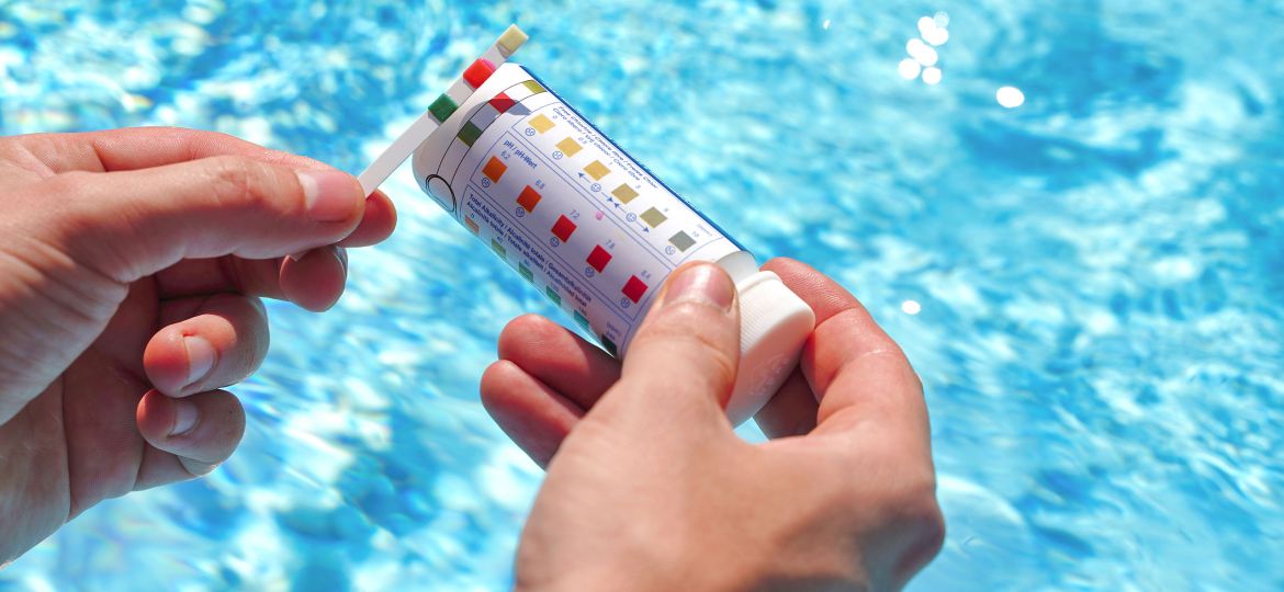 A person holds a pool test strip and a container with a color-coded chart in front of a sparkling blue swimming pool. The test strip is being compared to the chart, likely to assess water quality parameters such as pH and chlorine levels.