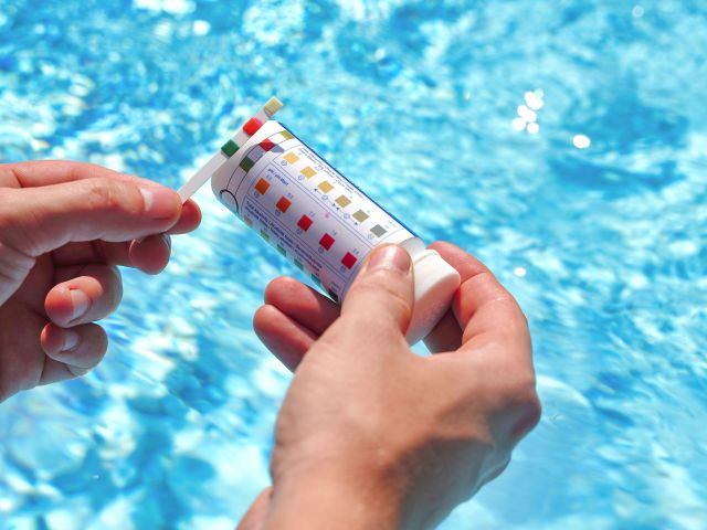 A person holds a pool test strip and a container with a color-coded chart in front of a sparkling blue swimming pool. The test strip is being compared to the chart, likely to assess water quality parameters such as pH and chlorine levels.