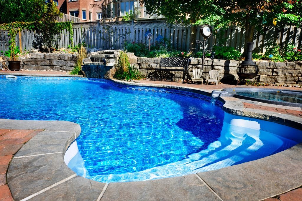 A backyard with a vibrant blue inground swimming pool surrounded by natural stone coping and red brick pavers. A small waterfall feature flows into the pool from a rock wall landscaped with plants. Adjacent to the pool is a circular hot tub with a stone edge and small water spillover. In the background, there is a wooden fence, a metal bench, two small Adirondack chairs, a vintage-style outdoor clock, and a chiminea fireplace among trees and shrubbery. Residential houses are partially visible beyond the fence.
