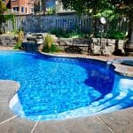 A backyard with a vibrant blue inground swimming pool surrounded by natural stone coping and red brick pavers. A small waterfall feature flows into the pool from a rock wall landscaped with plants. Adjacent to the pool is a circular hot tub with a stone edge and small water spillover. In the background, there is a wooden fence, a metal bench, two small Adirondack chairs, a vintage-style outdoor clock, and a chiminea fireplace among trees and shrubbery. Residential houses are partially visible beyond the fence.