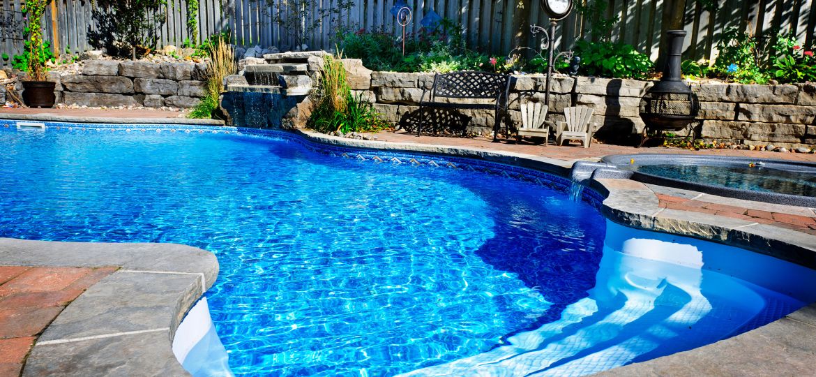 A backyard with a vibrant blue inground swimming pool surrounded by natural stone coping and red brick pavers. A small waterfall feature flows into the pool from a rock wall landscaped with plants. Adjacent to the pool is a circular hot tub with a stone edge and small water spillover. In the background, there is a wooden fence, a metal bench, two small Adirondack chairs, a vintage-style outdoor clock, and a chiminea fireplace among trees and shrubbery. Residential houses are partially visible beyond the fence.