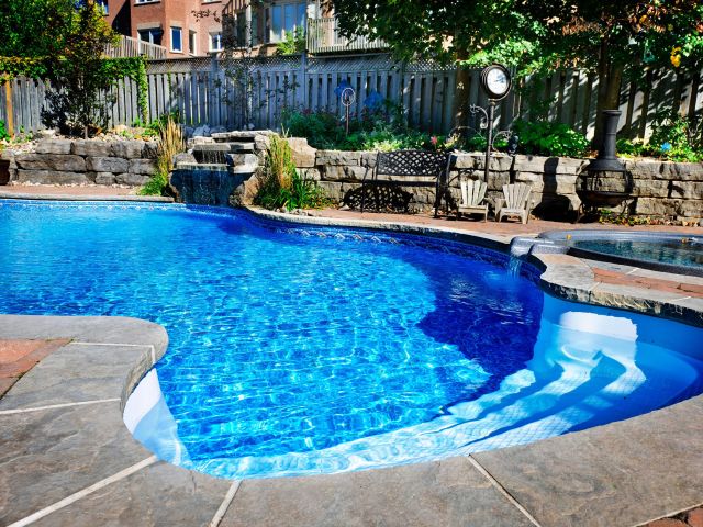 A backyard with a vibrant blue inground swimming pool surrounded by natural stone coping and red brick pavers. A small waterfall feature flows into the pool from a rock wall landscaped with plants. Adjacent to the pool is a circular hot tub with a stone edge and small water spillover. In the background, there is a wooden fence, a metal bench, two small Adirondack chairs, a vintage-style outdoor clock, and a chiminea fireplace among trees and shrubbery. Residential houses are partially visible beyond the fence.