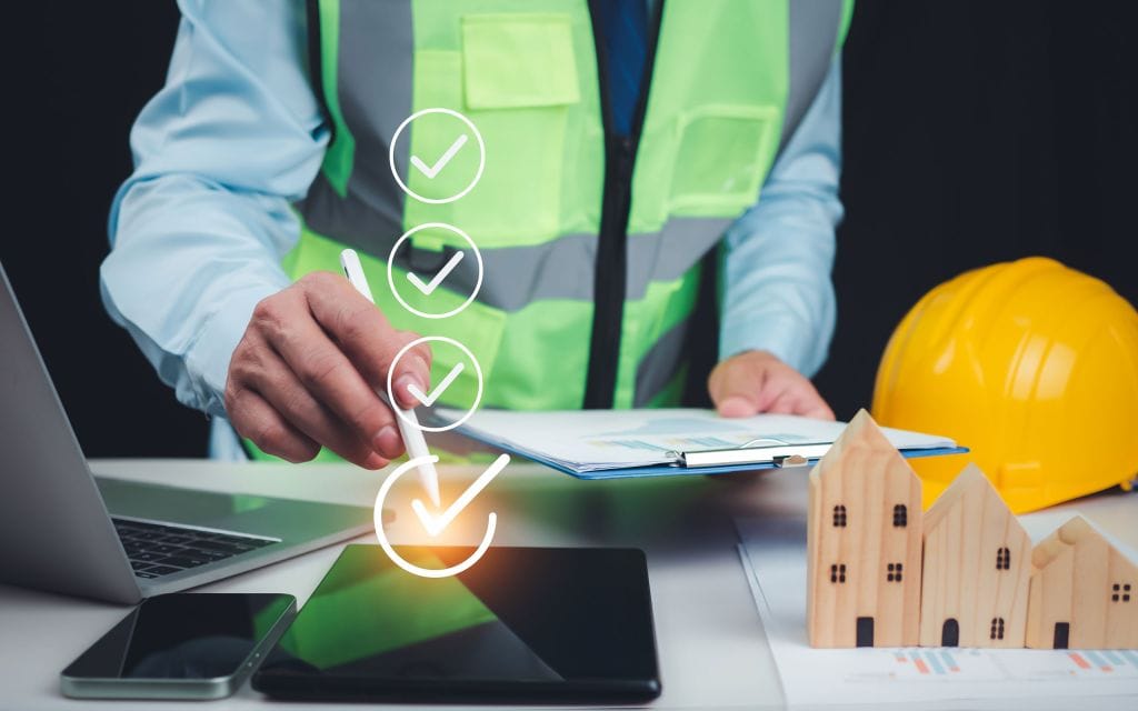 Construction project manager in a reflective safety vest reviewing documents on a clipboard, with a checklist graphic overlay and a stylus pointing to a digital tablet. Nearby are a laptop, smartphone, yellow hard hat, and wooden house models on a desk with charts.