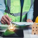 Construction project manager in a reflective safety vest reviewing documents on a clipboard, with a checklist graphic overlay and a stylus pointing to a digital tablet. Nearby are a laptop, smartphone, yellow hard hat, and wooden house models on a desk with charts.