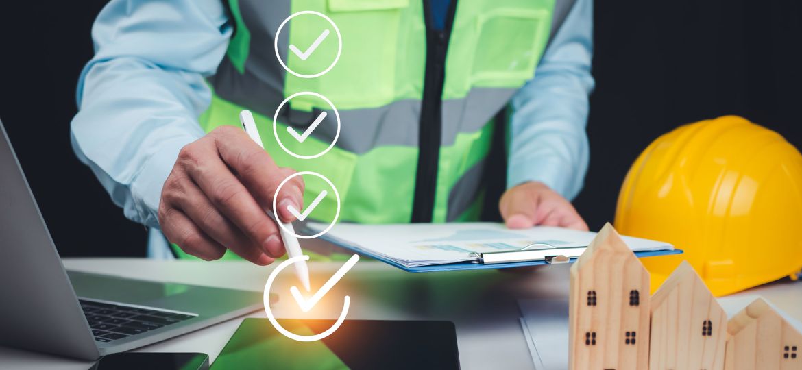 Construction project manager in a reflective safety vest reviewing documents on a clipboard, with a checklist graphic overlay and a stylus pointing to a digital tablet. Nearby are a laptop, smartphone, yellow hard hat, and wooden house models on a desk with charts.