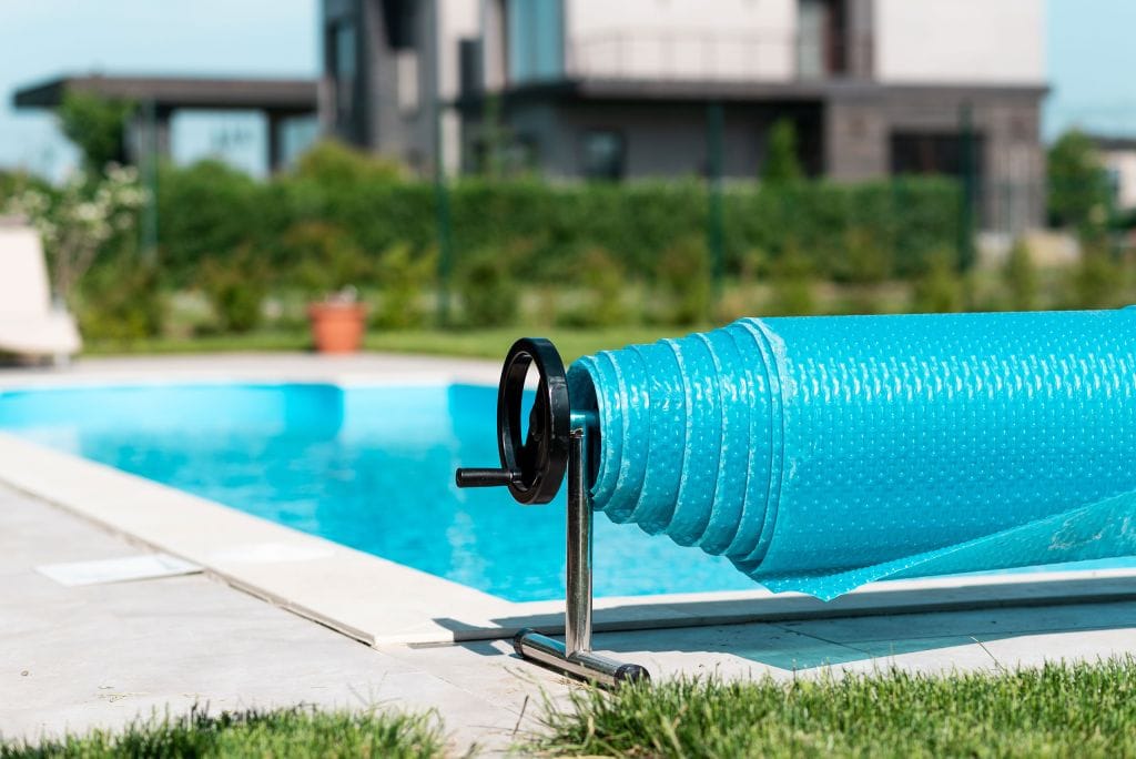 Close-up of a rolled-up blue bubble pool cover on a metal reel beside a clean outdoor swimming pool, with a modern house and green foliage in the background on a sunny day.