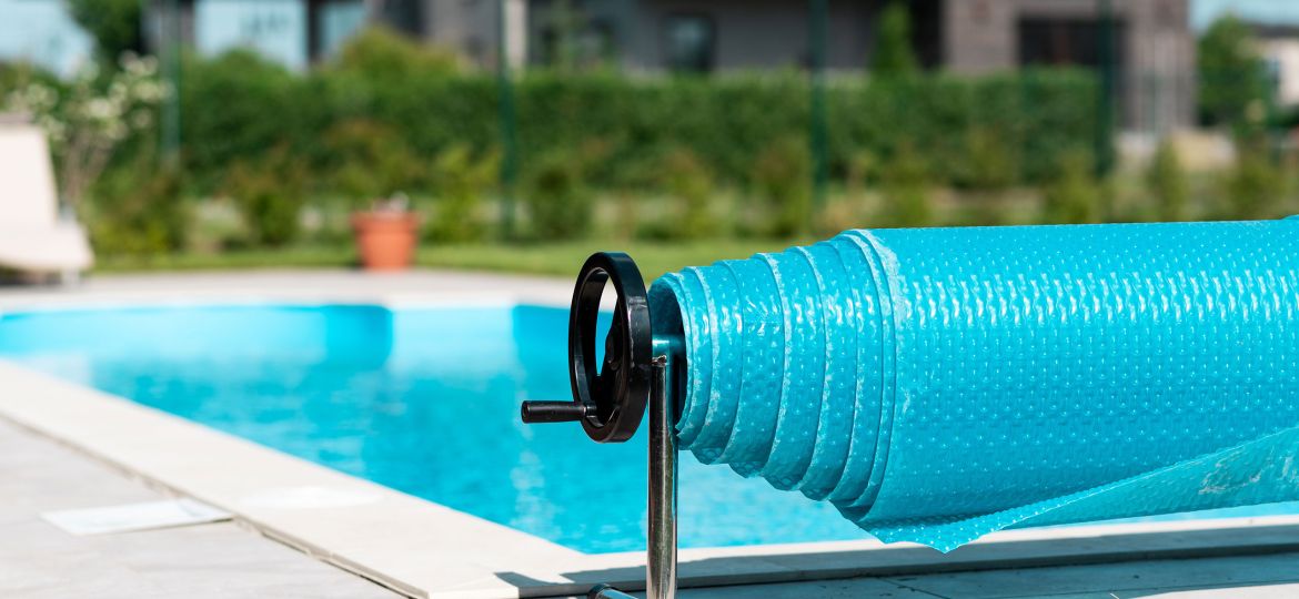 Close-up of a rolled-up blue bubble pool cover on a metal reel beside a clean outdoor swimming pool, with a modern house and green foliage in the background on a sunny day.