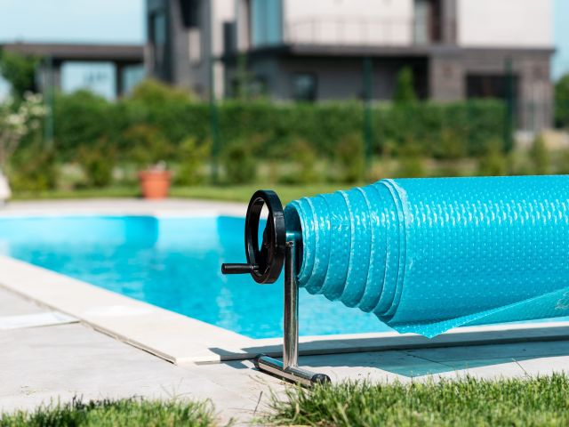 Close-up of a rolled-up blue bubble pool cover on a metal reel beside a clean outdoor swimming pool, with a modern house and green foliage in the background on a sunny day.