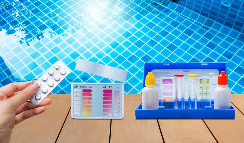 Hand holding a blister pack of pool test tablets near a poolside water testing kit. The kit includes color-coded charts for pH and chlorine levels, reagent bottles with red and yellow caps, and clear testing tubes, all set against the backdrop of a clear blue tiled swimming pool.