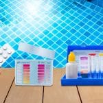 Hand holding a blister pack of pool test tablets near a poolside water testing kit. The kit includes color-coded charts for pH and chlorine levels, reagent bottles with red and yellow caps, and clear testing tubes, all set against the backdrop of a clear blue tiled swimming pool.