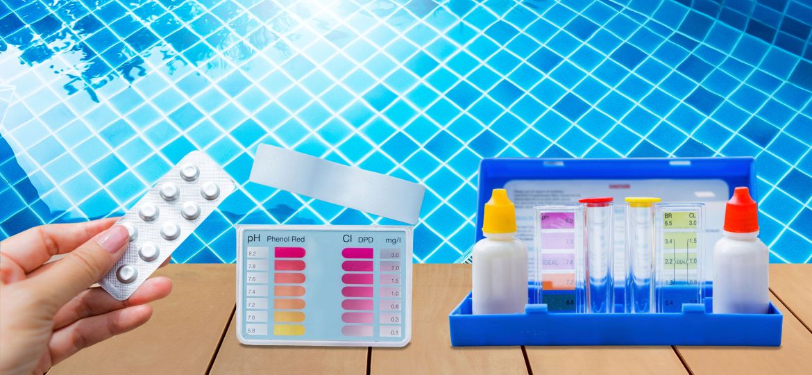 Hand holding a blister pack of pool test tablets near a poolside water testing kit. The kit includes color-coded charts for pH and chlorine levels, reagent bottles with red and yellow caps, and clear testing tubes, all set against the backdrop of a clear blue tiled swimming pool.