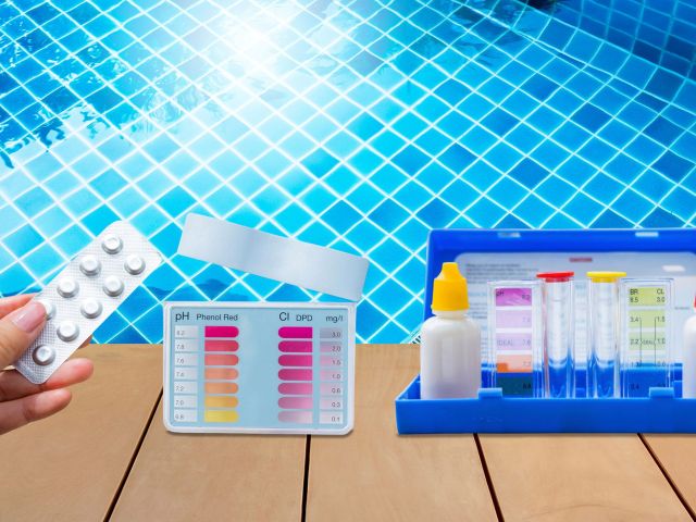 Hand holding a blister pack of pool test tablets near a poolside water testing kit. The kit includes color-coded charts for pH and chlorine levels, reagent bottles with red and yellow caps, and clear testing tubes, all set against the backdrop of a clear blue tiled swimming pool.