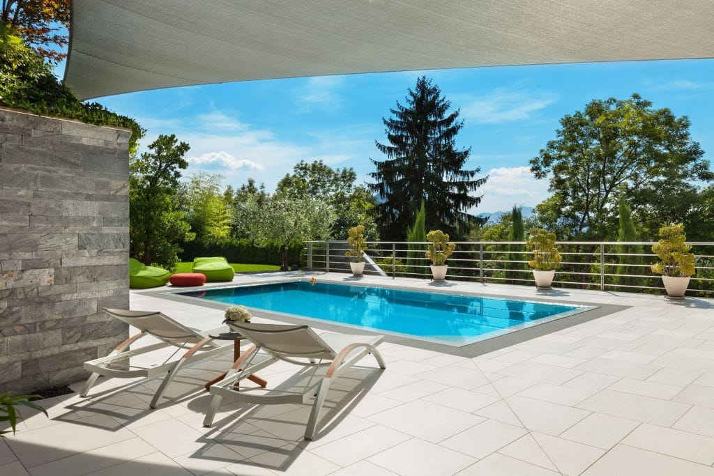 Modern outdoor pool area with white tiled flooring, two white sun loungers facing a rectangular blue swimming pool, and a small side table holding flowers. The pool is bordered by potted plants and surrounded by greenery, including tall trees and shrubs. Bright green bean bag chairs and a red ottoman sit near the pool, while a large shade sail stretches overhead against a clear blue sky.