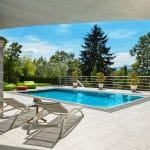 Modern outdoor pool area with white tiled flooring, two white sun loungers facing a rectangular blue swimming pool, and a small side table holding flowers. The pool is bordered by potted plants and surrounded by greenery, including tall trees and shrubs. Bright green bean bag chairs and a red ottoman sit near the pool, while a large shade sail stretches overhead against a clear blue sky.