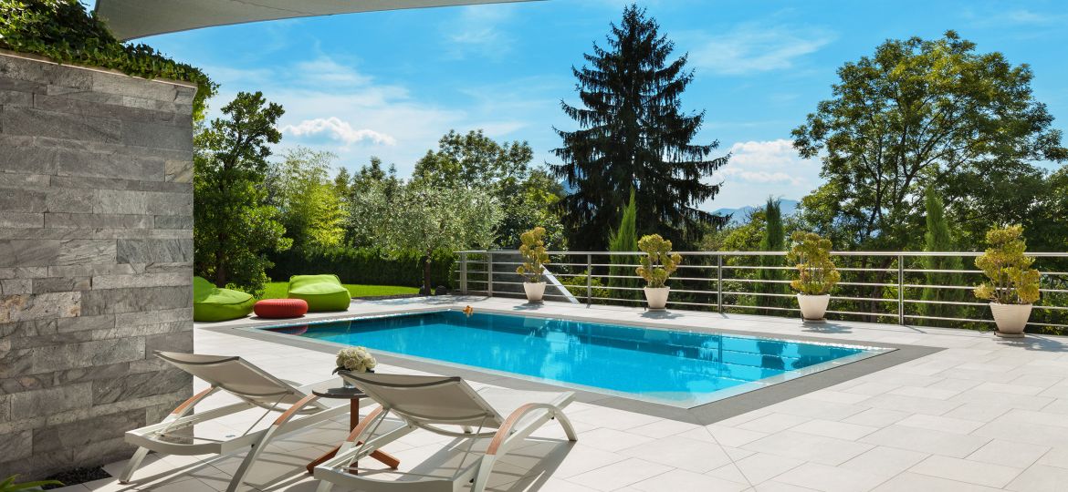 Modern outdoor pool area with white tiled flooring, two white sun loungers facing a rectangular blue swimming pool, and a small side table holding flowers. The pool is bordered by potted plants and surrounded by greenery, including tall trees and shrubs. Bright green bean bag chairs and a red ottoman sit near the pool, while a large shade sail stretches overhead against a clear blue sky.