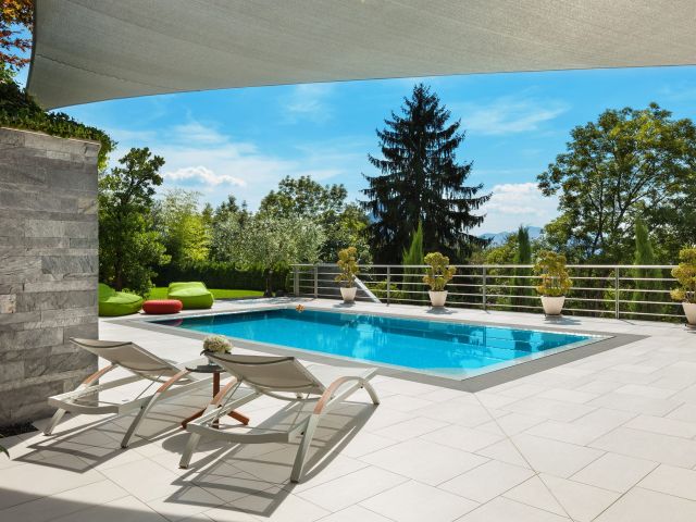 Modern outdoor pool area with white tiled flooring, two white sun loungers facing a rectangular blue swimming pool, and a small side table holding flowers. The pool is bordered by potted plants and surrounded by greenery, including tall trees and shrubs. Bright green bean bag chairs and a red ottoman sit near the pool, while a large shade sail stretches overhead against a clear blue sky.