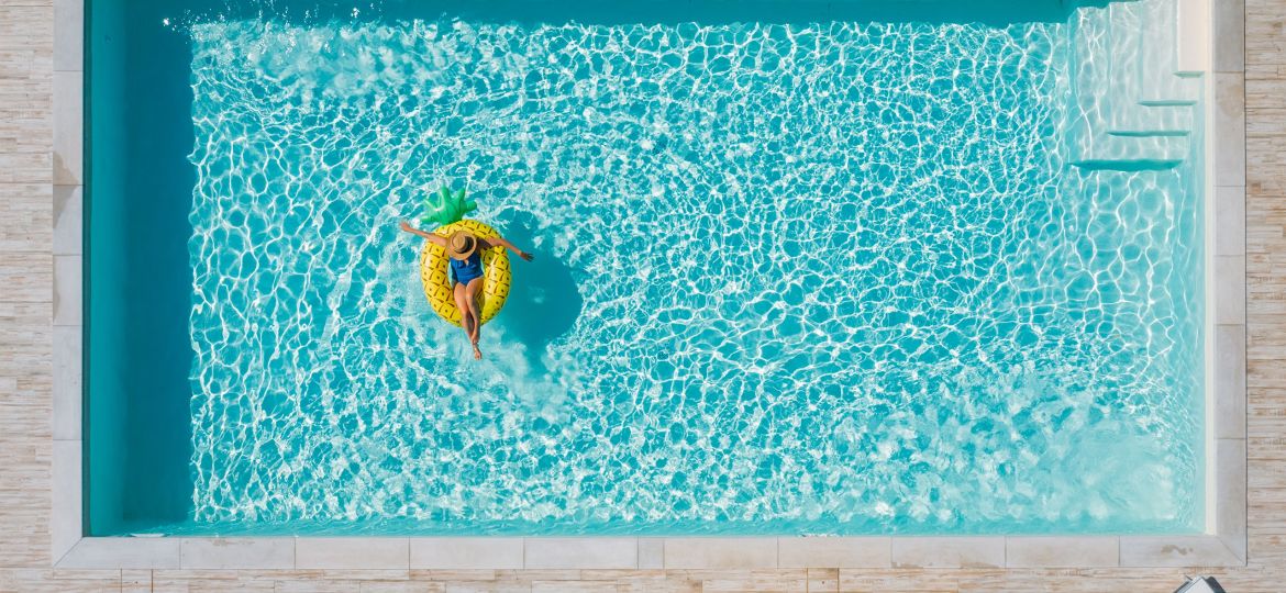 Person floating on a pineapple-shaped inflatable in a bright blue swimming pool, viewed from above.