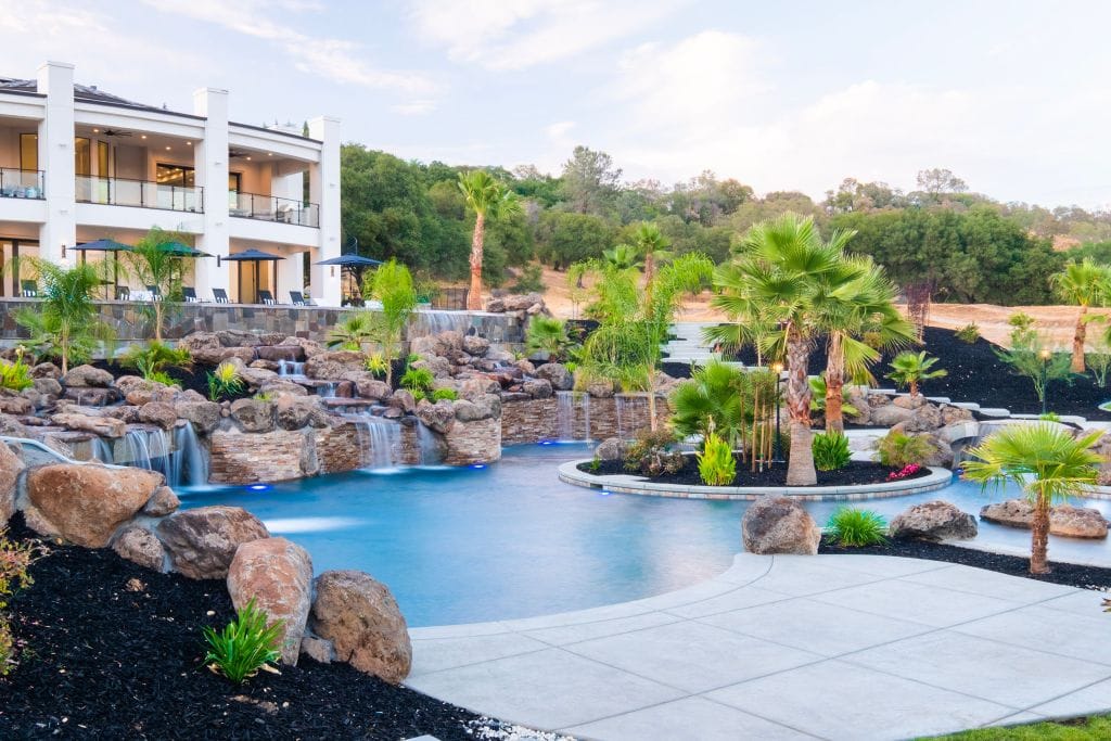 Luxury backyard pool with rock waterfalls, palm trees, and a modern white house in the background.