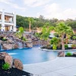 Luxury backyard pool with rock waterfalls, palm trees, and a modern white house in the background.