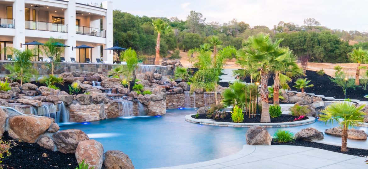 Luxury backyard pool with rock waterfalls, palm trees, and a modern white house in the background.