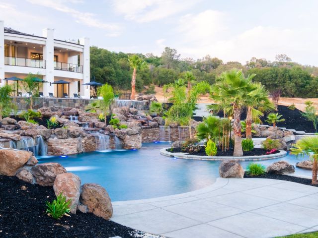 Luxury backyard pool with rock waterfalls, palm trees, and a modern white house in the background.