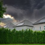 Houses with rooftop solar panels behind a tall green hedge under dark storm clouds at sunset.