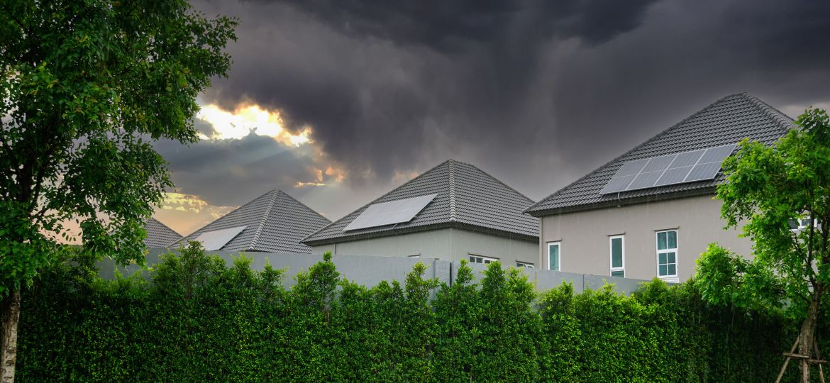 Houses with rooftop solar panels behind a tall green hedge under dark storm clouds at sunset.