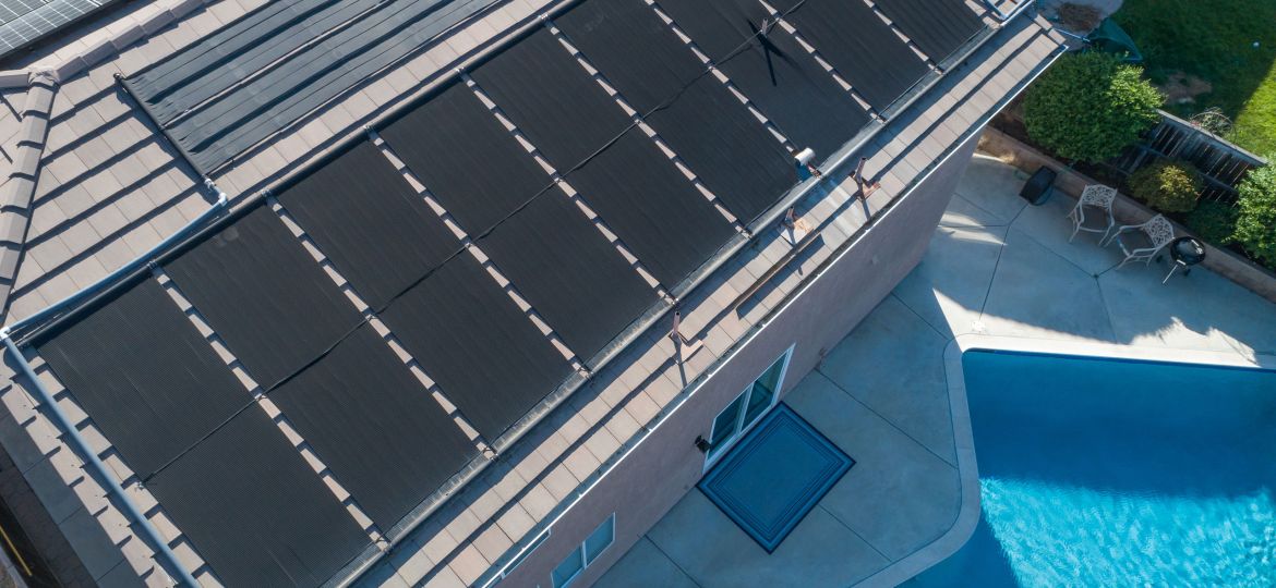 Black solar pool heating panels installed on a residential roof beside a backyard swimming pool.