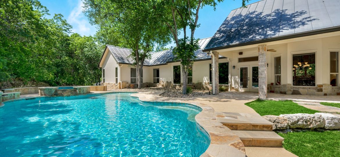 Curved backyard swimming pool beside a modern single-story home with metal roof and shaded patio