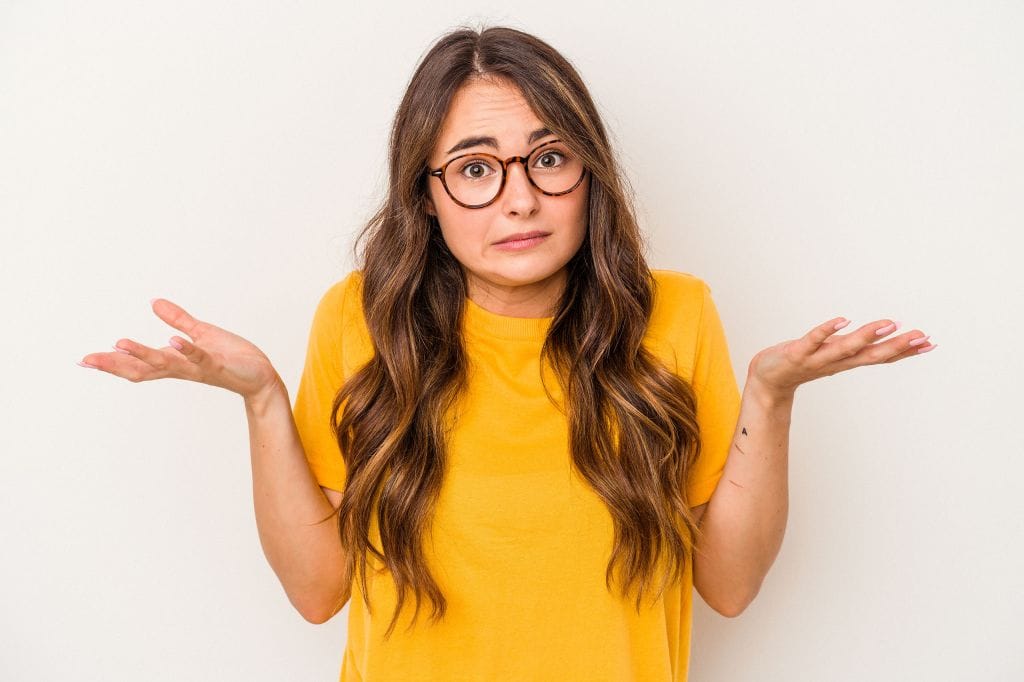 Woman in yellow shirt shrugging with raised hands and uncertain expression against a plain background